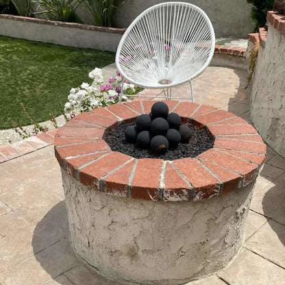 Small round stucco fire pit with a red brick cap, filled with black lava rock and a stacked cluster of Matte Black ceramic fire balls, set on a sunny patio near garden flowers and a white lounge chair.