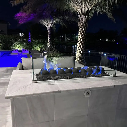 Nighttime poolside fire table with a glass wind guard, blue flames dancing over Matte Black 4-inch fire balls on black lava rock, with palm trees and a glowing blue pool in the background.