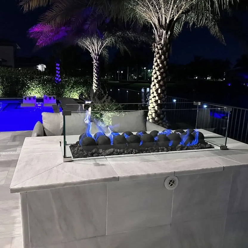 Nighttime poolside fire table with a glass wind guard, blue flames dancing over Matte Black 4-inch fire balls on black lava rock, with palm trees and a glowing blue pool in the background.