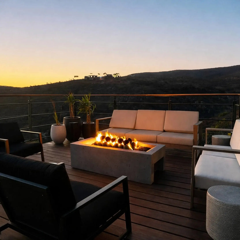 Modern rooftop-style deck at sunset with a rectangular concrete fire pit table burning around matte black fire balls, surrounded by outdoor lounge seating and a mountain view.