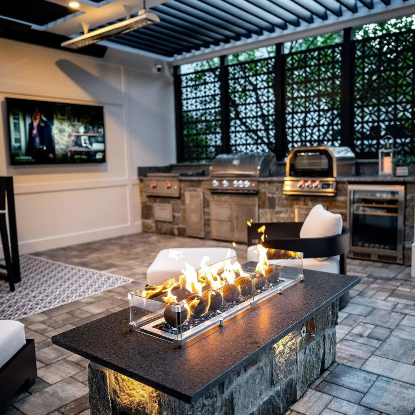 Covered patio outdoor kitchen with a stone fire table featuring a glass wind guard and tall flames rising over matte black fire balls, with built-in grills and paver flooring in the background.