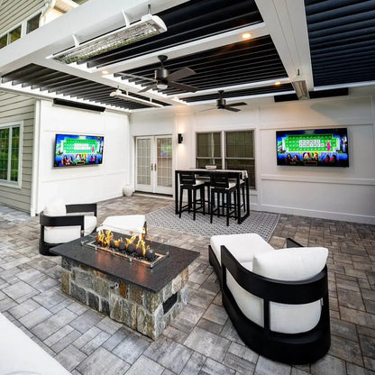 Wide view of a bright, modern covered patio with ceiling fans, TVs, bar seating, and a stone fire pit table where flames burn over matte black fire balls inside a clear glass guard.