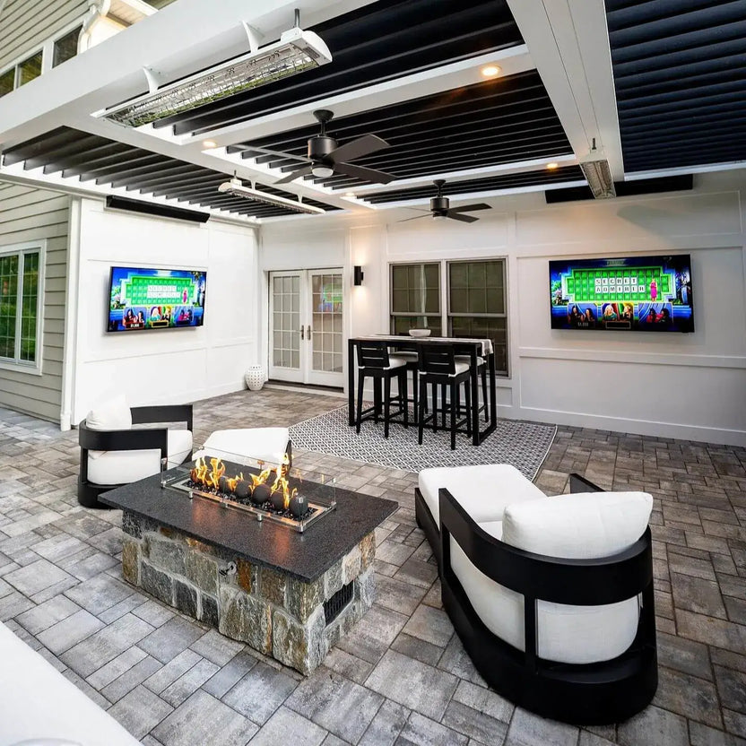 Wide view of a bright, modern covered patio with ceiling fans, TVs, bar seating, and a stone fire pit table where flames burn over matte black fire balls inside a clear glass guard.