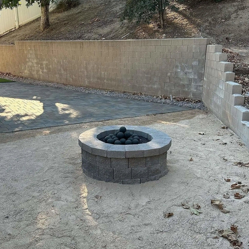 Backyard hardscape with a small round block fire pit filled with matte black fire balls, set on bare soil near a retaining wall and paved patio area.