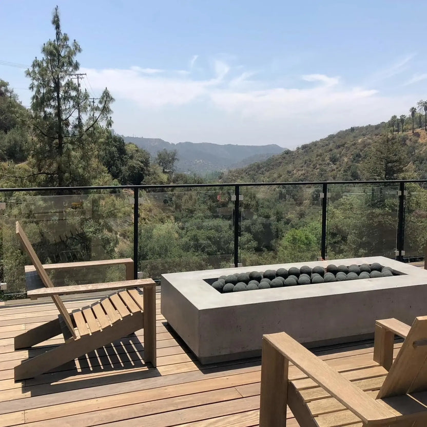 Contemporary rectangular concrete fire pit on a wood deck, filled edge-to-edge with Matte Black 4-inch fire balls in the burner, overlooking a scenic hillside view through a glass railing.