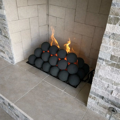 Close-up of a tiled fireplace corner showing stacked matte black fire balls on a burner tray, with yellow flames rising between the spheres against light gray firebrick.