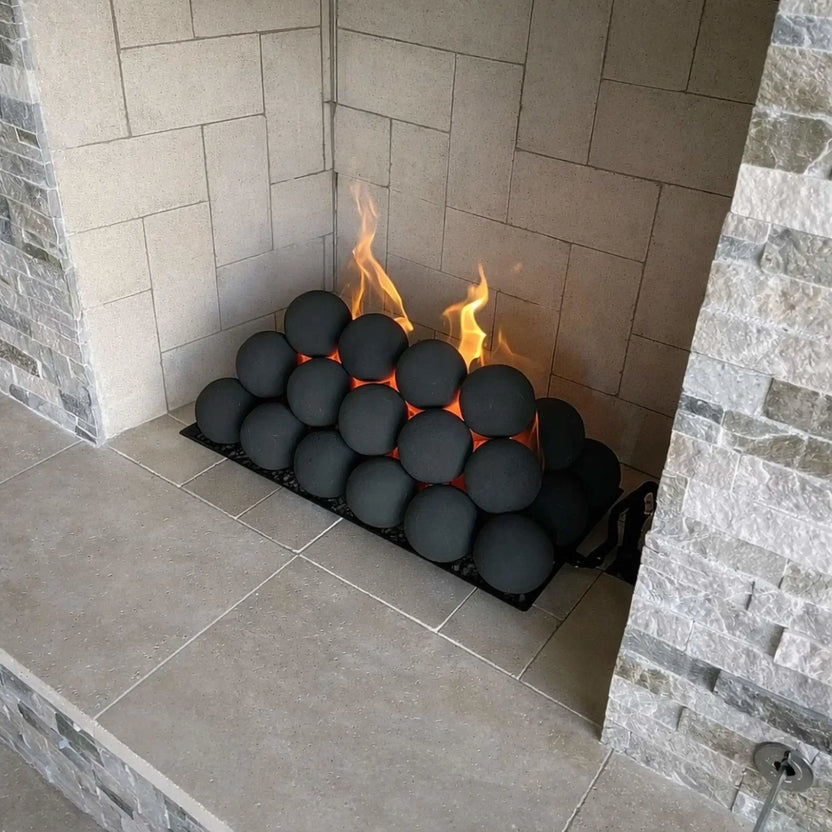 Close-up of a tiled fireplace corner showing stacked matte black fire balls on a burner tray, with yellow flames rising between the spheres against light gray firebrick.