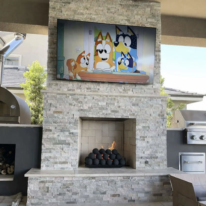 Outdoor stacked-stone fireplace with a wall-mounted TV above and a fireplace opening below, featuring matte black fire balls glowing with flames, flanked by patio grill stations.