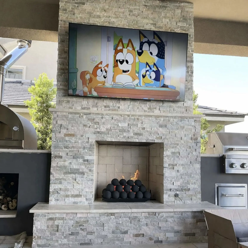 Outdoor stacked-stone fireplace with a wall-mounted TV above and a fireplace opening below, featuring matte black fire balls glowing with flames, flanked by patio grill stations.