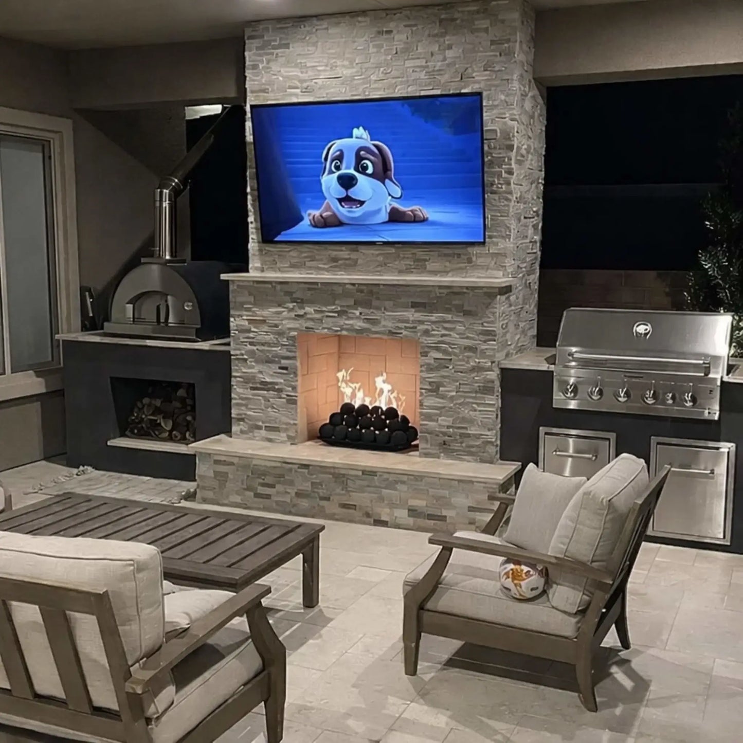 Nighttime outdoor living space with a stacked-stone fireplace and TV, where flames flicker over matte black fire balls inside the firebox, with patio seating and grills nearby.