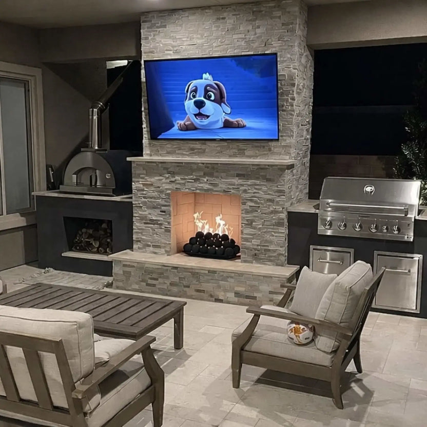Nighttime outdoor living space with a stacked-stone fireplace and TV, where flames flicker over matte black fire balls inside the firebox, with patio seating and grills nearby.