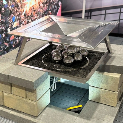 Rectangular fire pit table with a hinged stainless steel burner cover partially open, revealing polished stainless steel fire balls arranged over black lava rock inside the burner tray.