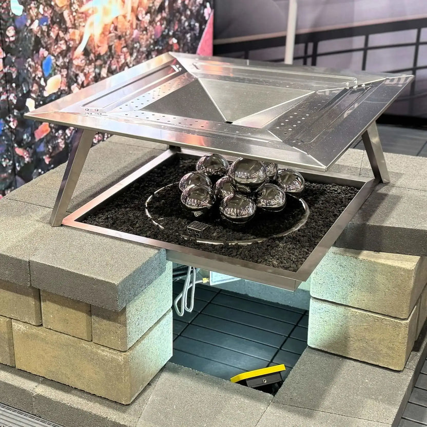 Rectangular fire pit table with a hinged stainless steel burner cover partially open, revealing polished stainless steel fire balls arranged over black lava rock inside the burner tray.