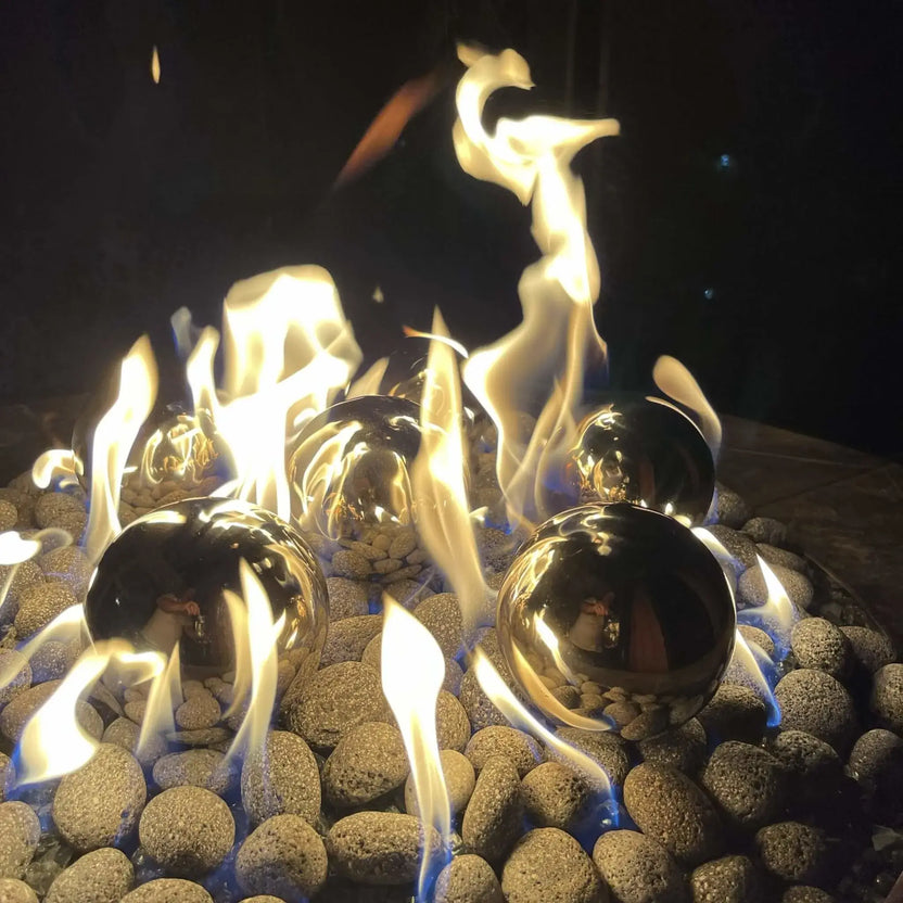 Nighttime close-up of stainless steel fire balls surrounded by tall yellow flames, reflecting the firelight while resting on rounded lava stones inside a gas fire pit.