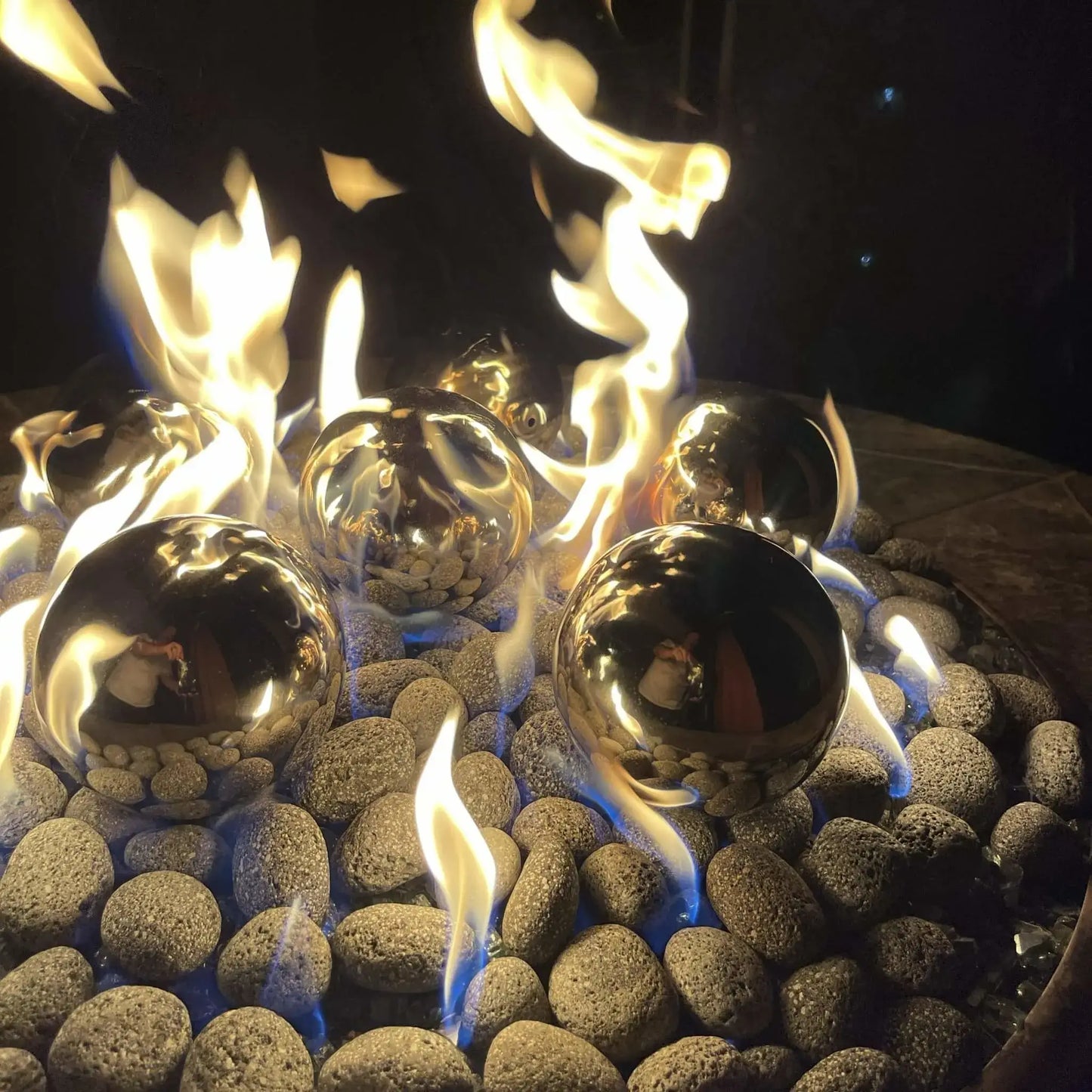 Stainless steel fire balls placed in a modern gas fire pit, reflecting dancing flames and surrounding stones, with blue and yellow fire burning between smooth river rock media for a contemporary outdoor fire feature.