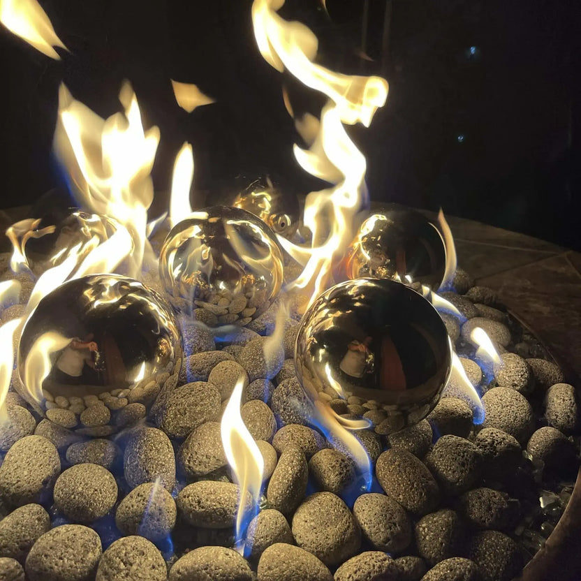 Stainless steel fire balls placed in a modern gas fire pit, reflecting dancing flames and surrounding stones, with blue and yellow fire burning between smooth river rock media for a contemporary outdoor fire feature.