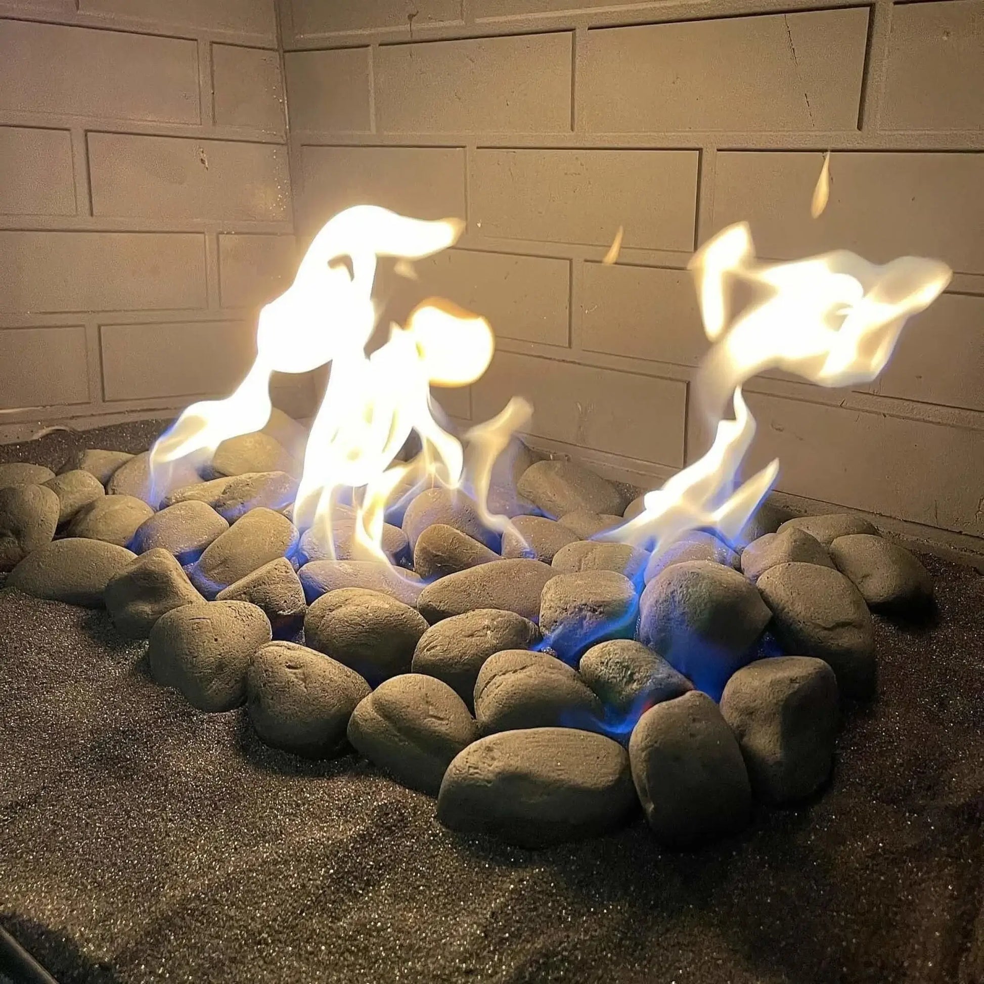 Close-up lifestyle view of American Fire Products Lite Stones in 15-inch Cape Gray arranged in a circular formation inside a gas fireplace, with smooth, rounded gray stones set over a dark sand base as natural yellow flames rise above blue gas flames, framed by a light brick interior wall for a clean, modern hearth look.