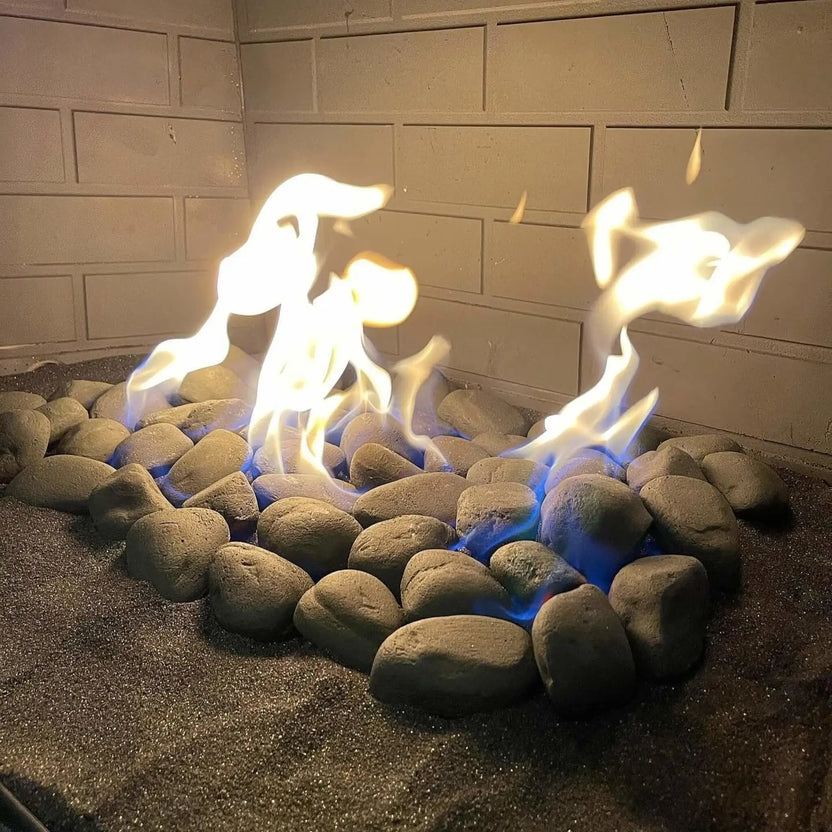 Close-up lifestyle view of American Fire Products Lite Stones in 15-inch Cape Gray arranged in a circular formation inside a gas fireplace, with smooth, rounded gray stones set over a dark sand base as natural yellow flames rise above blue gas flames, framed by a light brick interior wall for a clean, modern hearth look.