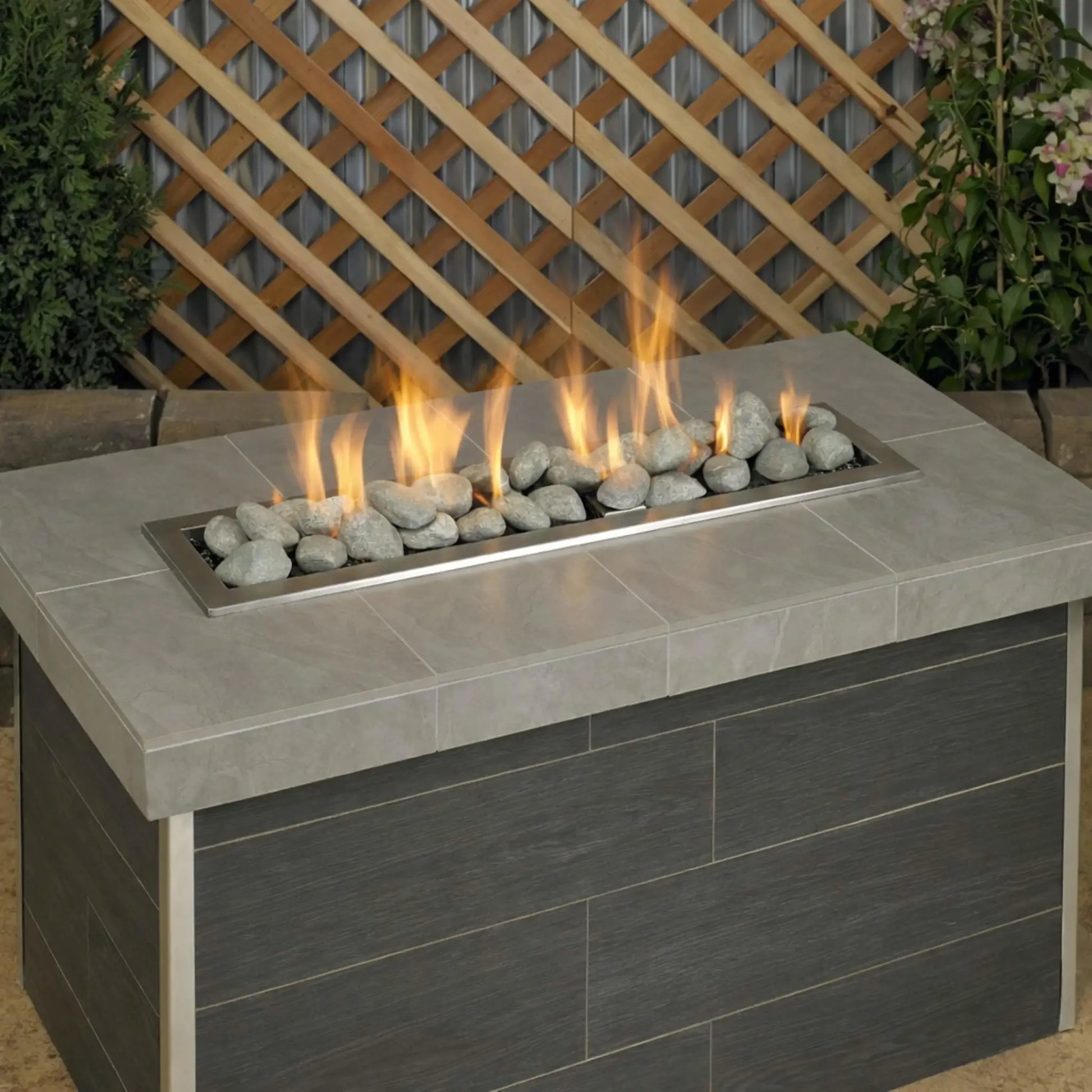 Rectangular outdoor gas fire table filled with Cape Gray Lite Stones by American Fire Products, featuring vibrant flames rising through smooth gray stones in a modern backyard patio setting.