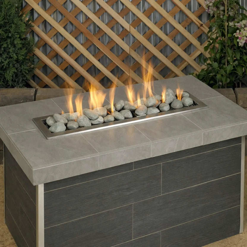 Rectangular outdoor gas fire table filled with Cape Gray Lite Stones by American Fire Products, featuring vibrant flames rising through smooth gray stones in a modern backyard patio setting.