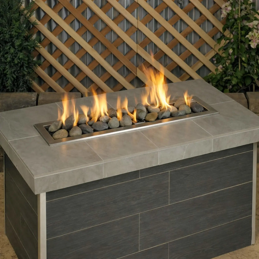 Outdoor linear gas fire table burning with bright flames over Natural Lite Stones by American Fire Products, showcasing a blend of earth-tone stones that create a realistic and organic fire pit appearance.