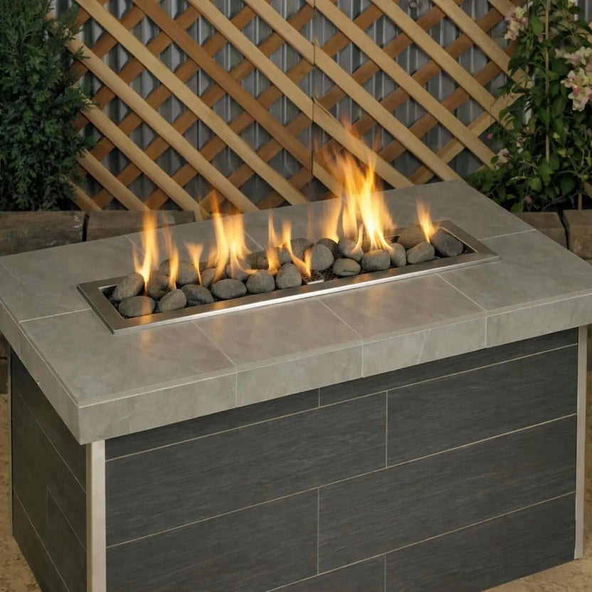 Modern outdoor linear gas fire table burning with tall yellow flames over Thunder Gray Lite Stones by American Fire Products, showcasing dark gray rounded stones set into a stainless steel burner for a contemporary patio design.
