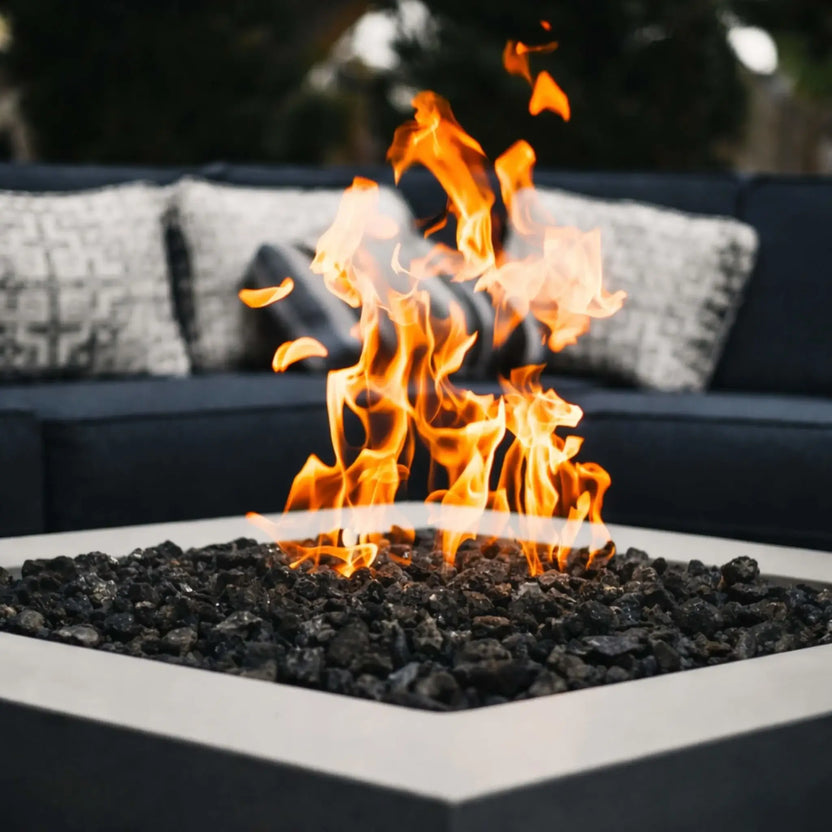 Modern square outdoor gas fire pit filled with black lava rock, producing bright orange flames for a clean, contemporary backyard patio setting.