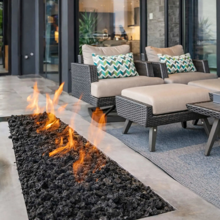 Contemporary linear gas fire pit installed beside patio seating, filled with black lava rock media that enhances flame height and heat distribution.