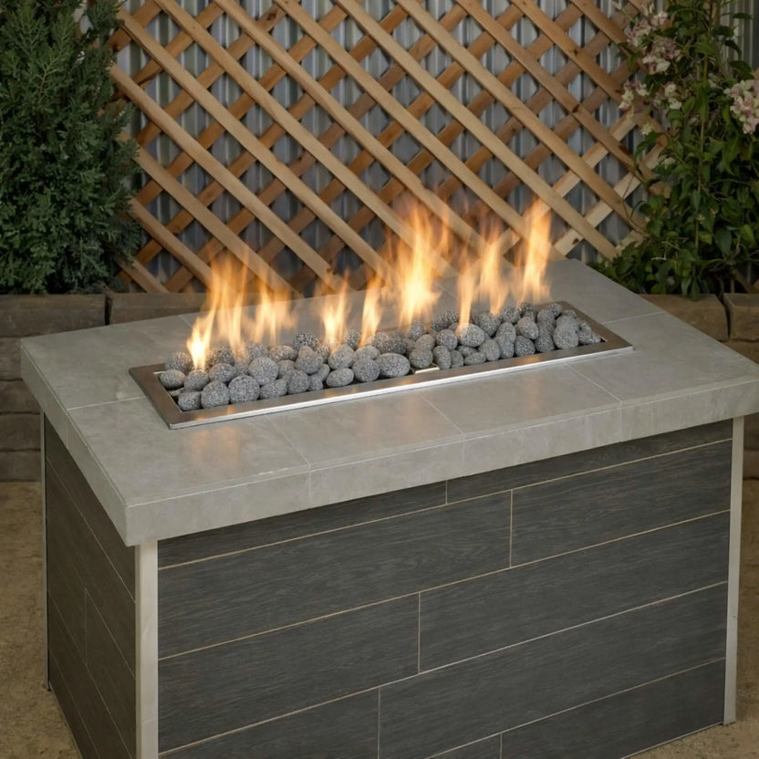 Contemporary rectangular outdoor fire table filled with medium lava stones lining a stainless steel linear burner, with steady yellow flames rising in front of a backyard wood lattice and garden plants.