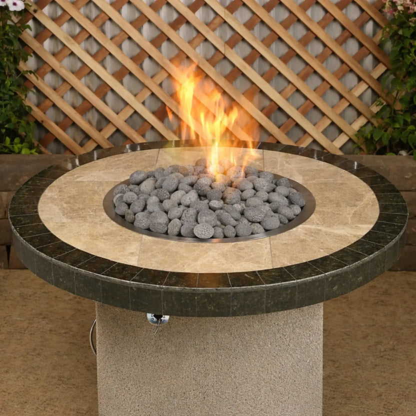 Round stone-top patio fire pit featuring medium gray lava rocks surrounding a central gas flame, set in an outdoor backyard space with lattice fencing and greenery.