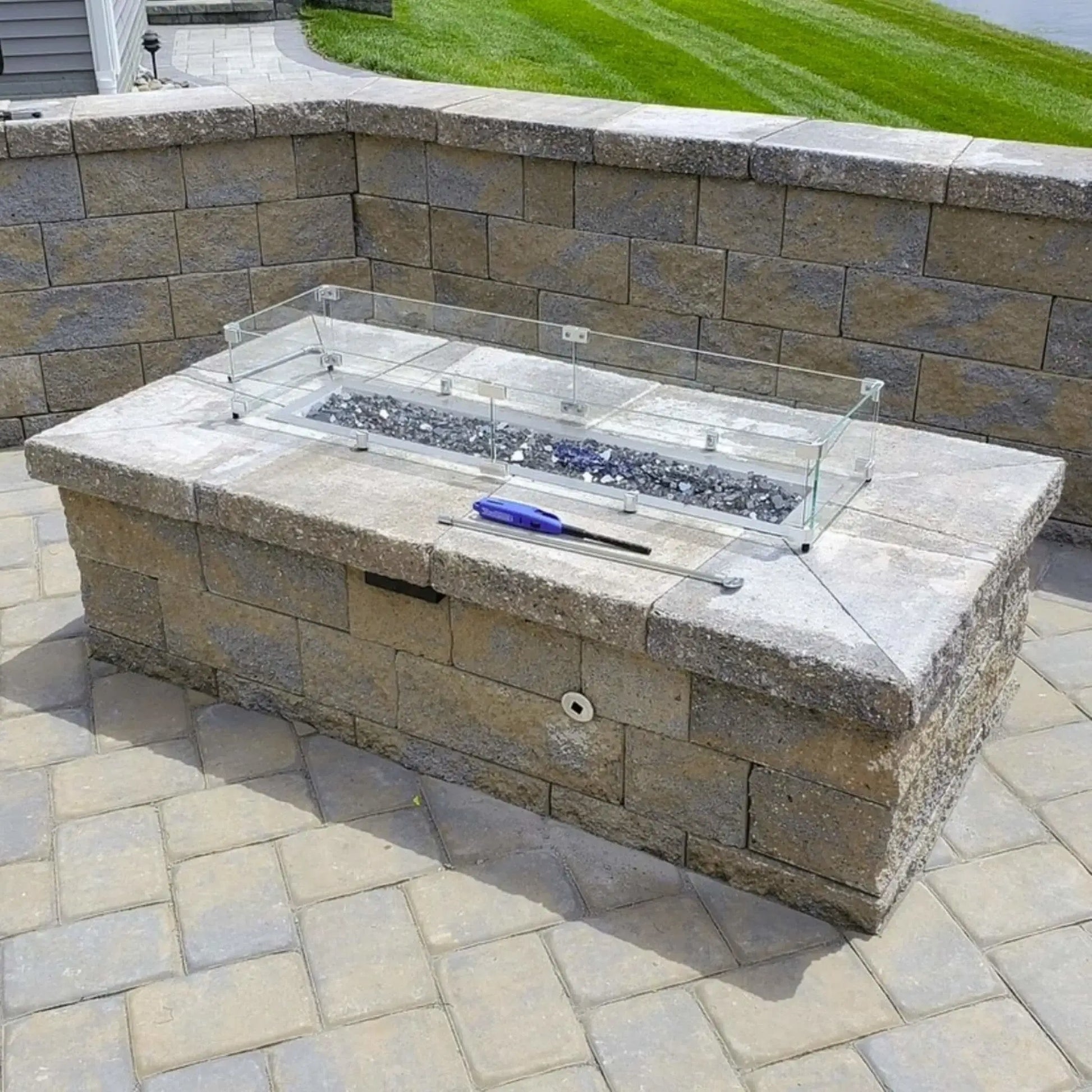 Built in stone fire pit with glass wind guard and fire glass media, photographed during installation, illustrating the use of a polished chrome universal gas key for operating the gas control valve.
