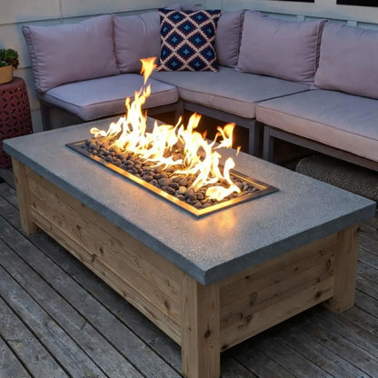 Rectangular outdoor fire pit table featuring a drop-in burner pan set into a concrete-style top, producing tall natural gas flames over river rock, surrounded by cushioned sectional seating on a wood deck.