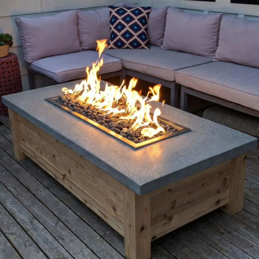Rectangular outdoor fire pit table featuring a drop-in burner pan set into a concrete-style top, producing tall natural gas flames over river rock, surrounded by cushioned sectional seating on a wood deck.