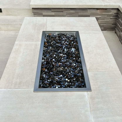 Top-down view of a rectangular drop-in burner pan installed in a concrete fire pit table, filled with reflective black fire glass and framed by smooth light-colored stone surfaces.