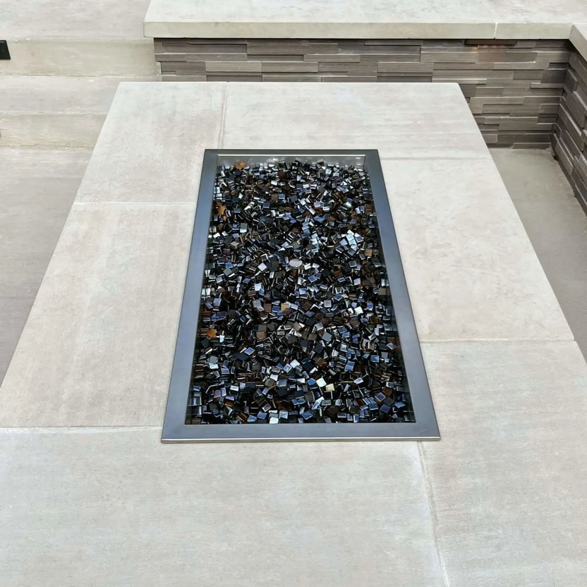 Top-down view of a rectangular drop-in burner pan installed in a concrete fire pit table, filled with reflective black fire glass and framed by smooth light-colored stone surfaces.