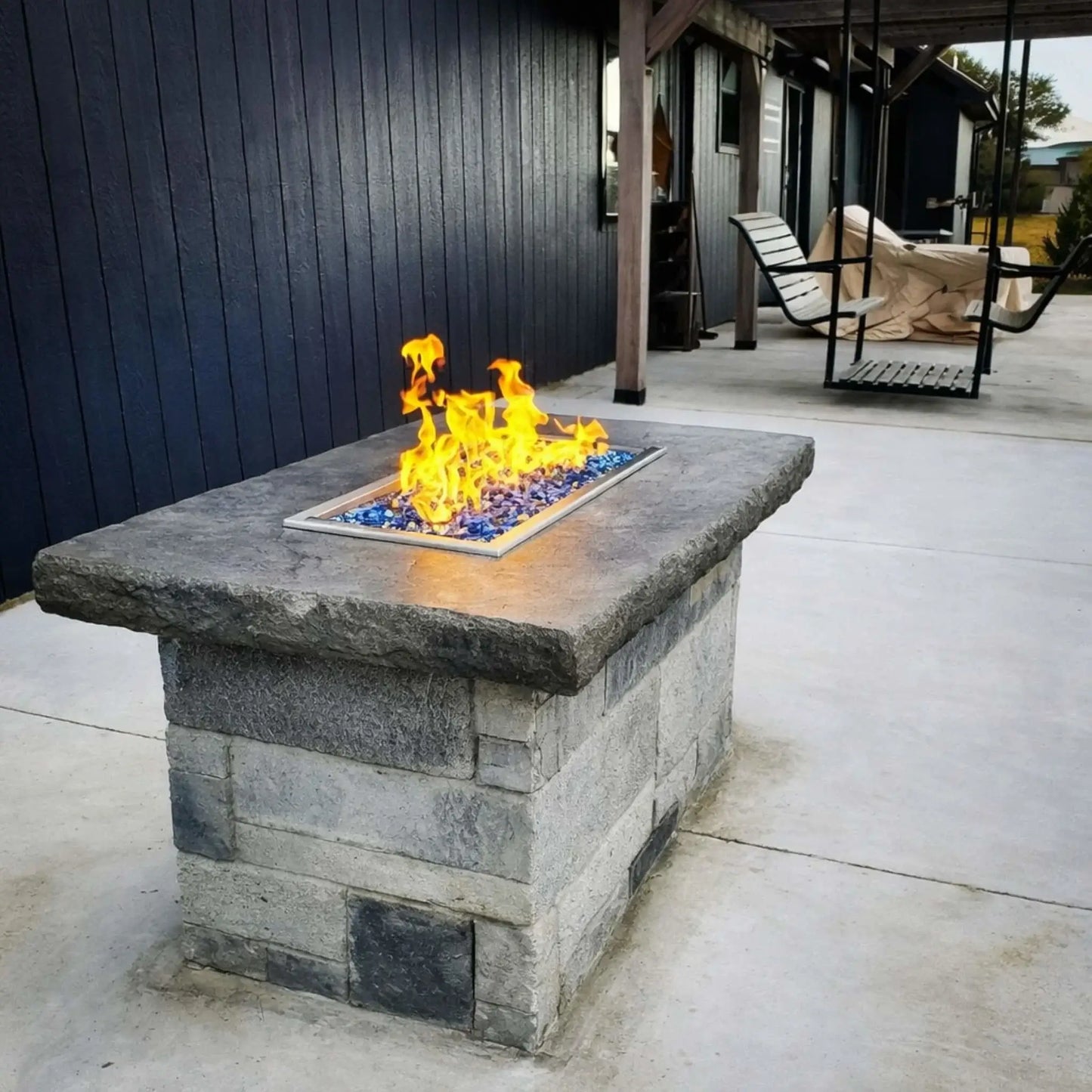 Rectangular stone fire pit with a drop-in burner pan producing yellow flames over blue fire glass, positioned on a concrete patio beside a covered seating area with hanging swing chairs.