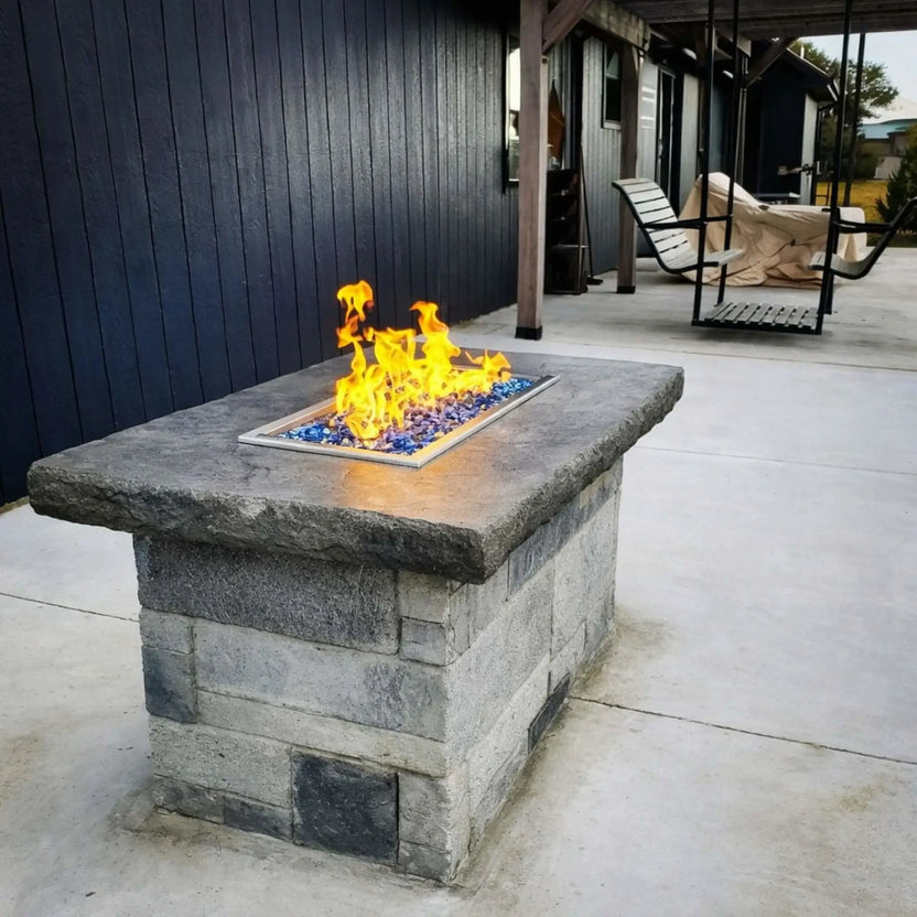 Rectangular stone fire pit with a drop-in burner pan producing yellow flames over blue fire glass, positioned on a concrete patio beside a covered seating area with hanging swing chairs.