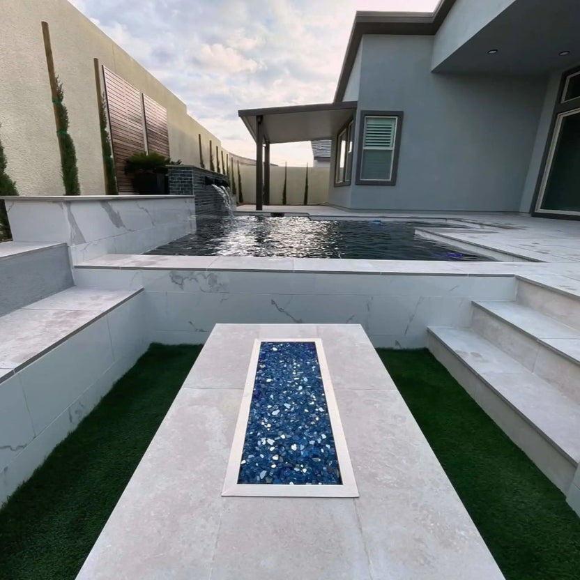 Modern poolside fire feature showcasing a rectangular drop-in burner pan filled with blue fire glass, installed flush into a raised stone platform overlooking a swimming pool with water features and stepped seating.