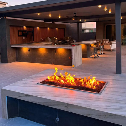 Upscale outdoor living space with a large covered kitchen and bar area, highlighted by a rectangular fire pit table featuring a drop-in burner pan with active flames set into a marble-style stone surface.