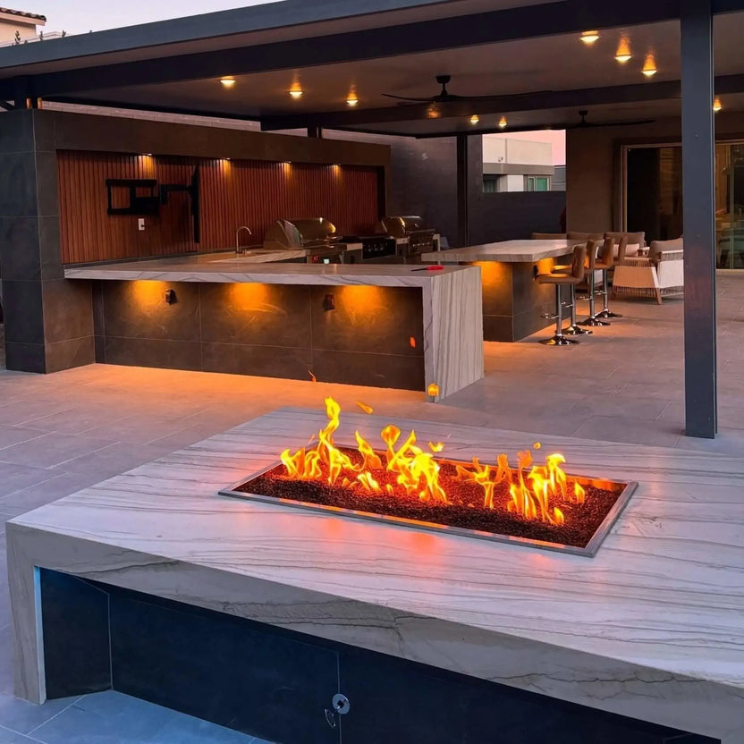 Upscale outdoor living space with a large covered kitchen and bar area, highlighted by a rectangular fire pit table featuring a drop-in burner pan with active flames set into a marble-style stone surface.