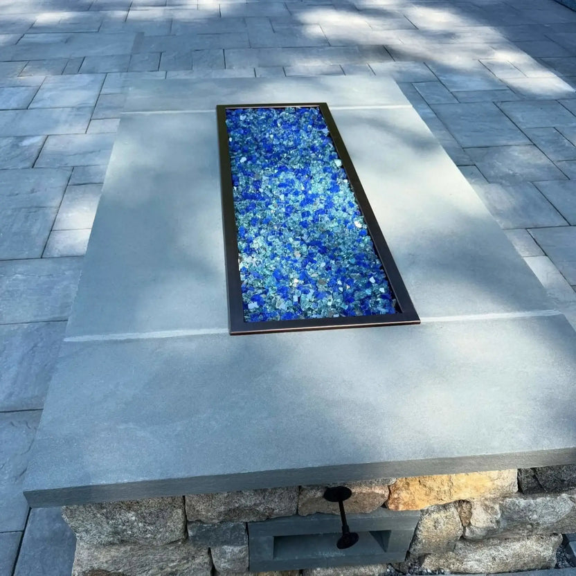 Rectangular outdoor fire pit table with a smooth gray stone top, featuring a drop-in burner pan filled with blue fire glass, set into a raised stone base on a paved patio with soft natural light and tree shadows.