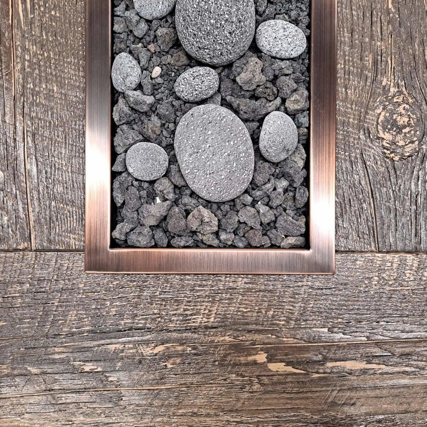 Close-up view of a rectangular drop-in burner pan with an oil-rubbed bronze trim, filled with lava rock and smooth decorative river stones, installed flush into a rustic wood-style fire table surface.