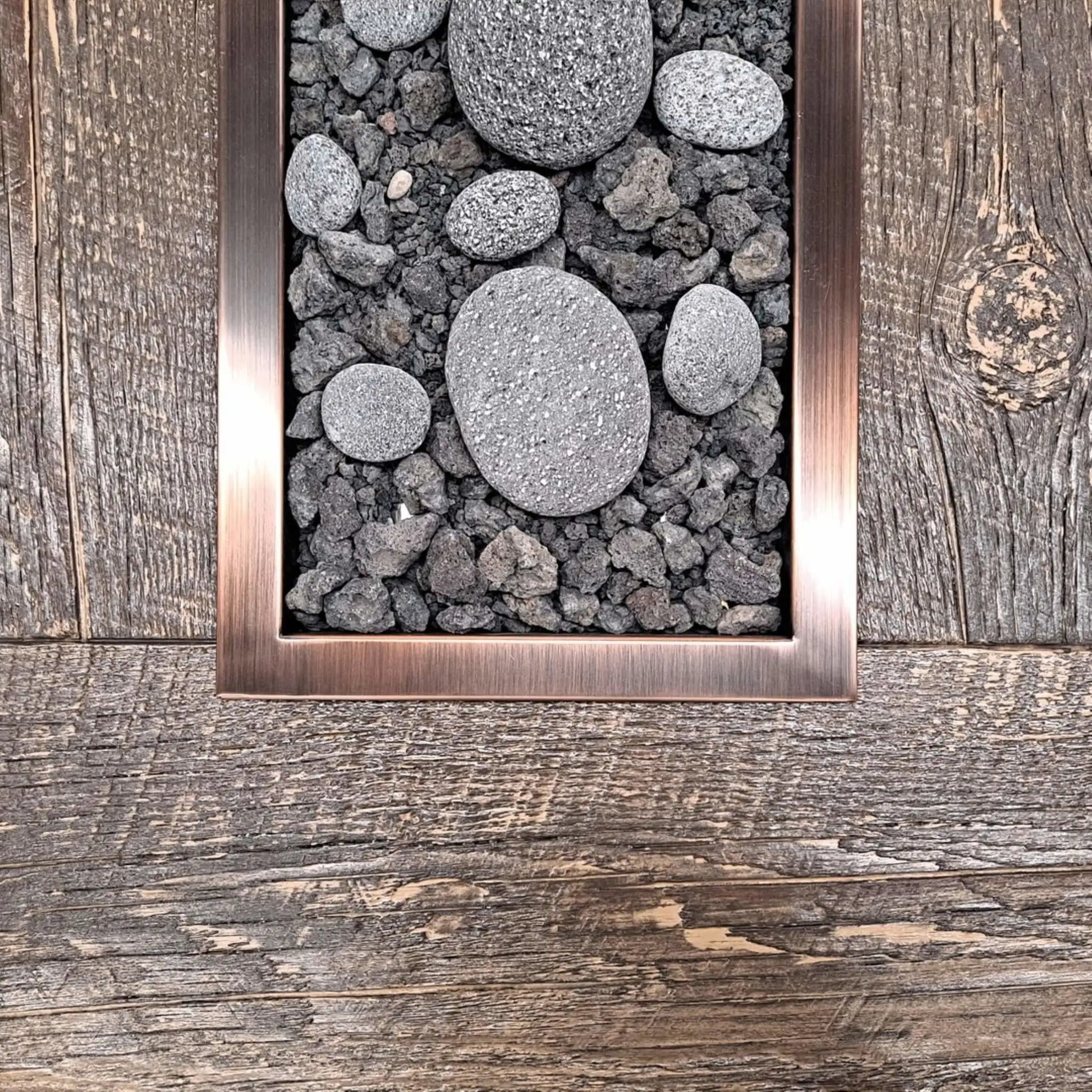 Close-up view of a rectangular drop-in burner pan with an oil-rubbed bronze trim, filled with lava rock and smooth decorative river stones, installed flush into a rustic wood-style fire table surface.