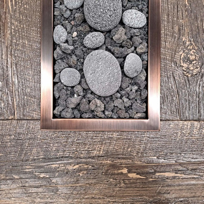 Close-up view of a rectangular drop-in burner pan with an oil-rubbed bronze trim, filled with lava rock and smooth decorative river stones, installed flush into a rustic wood-style fire table surface.