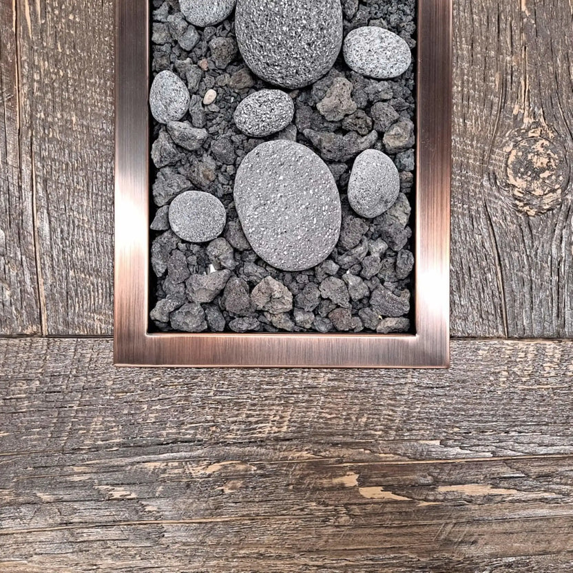 Close-up view of a rectangular drop-in burner pan with an oil-rubbed bronze trim, filled with lava rock and smooth decorative river stones, installed flush into a rustic wood-style fire table surface.