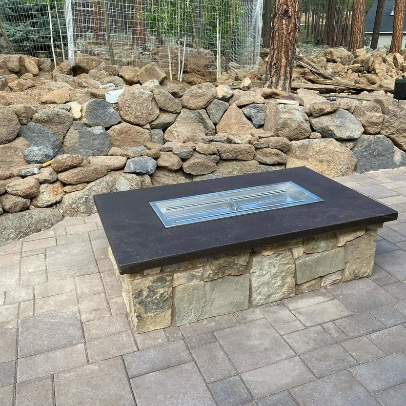 Custom rectangular fire pit table with a dark stone countertop and stainless steel drop-in burner pan installed, shown without fire media, set on a paved patio surrounded by natural stone retaining walls and trees.