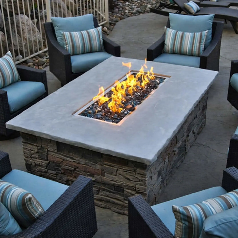 Outdoor seating area featuring a rectangular gas fire pit table with stacked stone sides and a smooth concrete top, showcasing a drop-in burner pan with black fire glass and active flames surrounded by cushioned patio chairs.