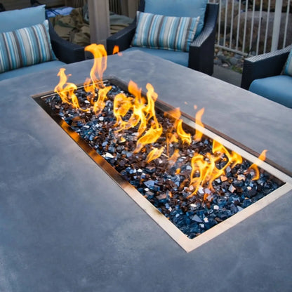 Detailed angled view of a rectangular drop-in burner pan producing bright yellow flames over reflective fire glass, set into a gray concrete fire pit table within a cozy outdoor lounge seating arrangement.