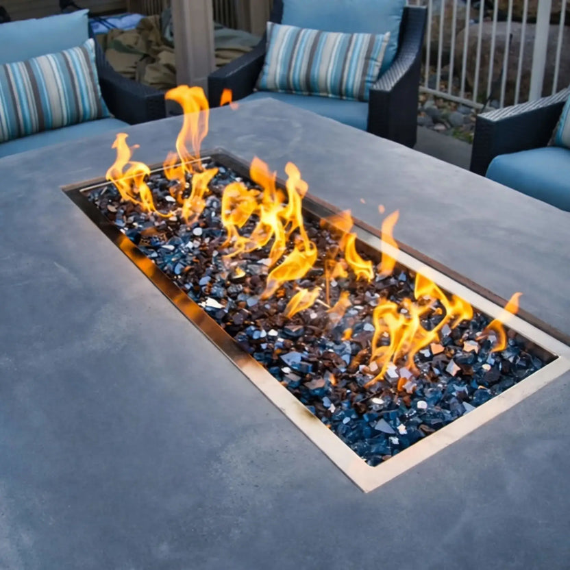 Detailed angled view of a rectangular drop-in burner pan producing bright yellow flames over reflective fire glass, set into a gray concrete fire pit table within a cozy outdoor lounge seating arrangement.