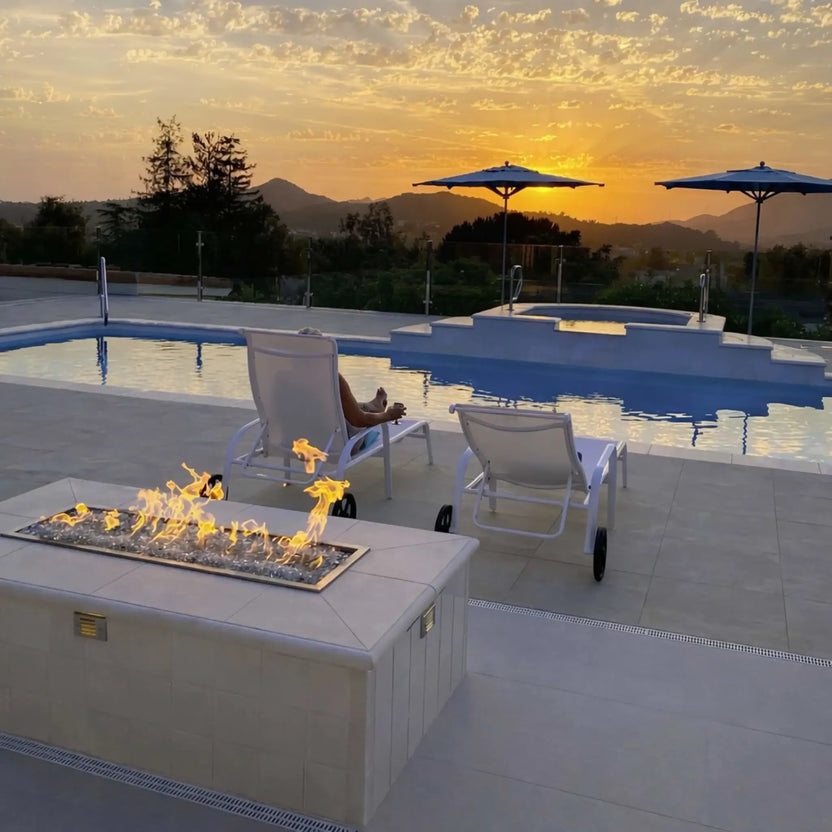 Poolside outdoor fire feature with a rectangular drop-in burner pan installed in a tiled fire pit, showcasing linear flames over fire glass with lounge chairs and a sunset mountain view in the background.